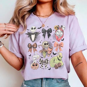 K&ouml;nnte beinhalten: Ein helllila T-Shirt mit einem Design mit Charakteren und Schleifen aus The Nightmare Before Christmas. Die Grafik zeigt Jack Skellington, Sally und Oogie Boogie mit Spinnweben und Flederm&auml;usen.