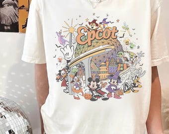 Puede incluir: Una camiseta de manga corta color crema con un estampado gráfico colorido de varios personajes de dibujos animados con disfraces de Halloween, la esfera geodésica de Epcot, un monorraíl y el texto Epcot en una fuente naranja estilizada.