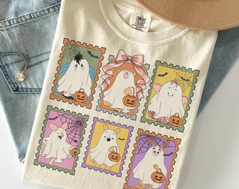Puede incluir: Una camiseta de color crema con un estampado gráfico de seis ilustraciones estilo sello postal. Cada sello muestra a un personaje de dibujos animados disfrazado de fantasma, sosteniendo una calabaza, con telarañas y murciélagos.