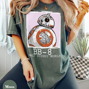 May include: Moss green t-shirt with a BB-8 droid graphic from Star Wars, set on a pink background. The text "BB-8 WALT DISNEY WORLD" is printed below. The shirt has a relaxed fit.