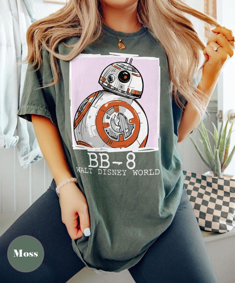 May include: Moss green t-shirt with a BB-8 droid graphic from Star Wars, set on a pink background. The text "BB-8 WALT DISNEY WORLD" is printed below. The shirt has a relaxed fit.