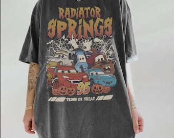 Puede incluir: Una camiseta gris carbón de manga corta con un estampado gráfico que incluye el texto Radiator Springs y Trunk or Treat. El diseño muestra personajes de coches, telarañas, calabazas y el número 95 en tonos naranja y amarillo.