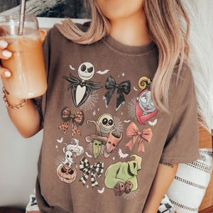 K&ouml;nnte beinhalten: Ein braunes T-Shirt mit Cartoon-Charakteren und Schleifen. Das Design umfasst Jack Skellington, Sally und Oogie Boogie, zusammen mit Flederm&auml;usen und Spinnweben. Die Schleifen sind in verschiedenen Farben und Mustern.