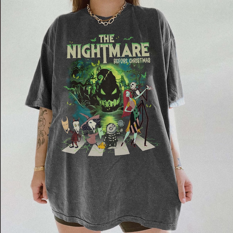 May include: A charcoal gray short-sleeve t-shirt featuring a graphic print of The Nightmare Before Christmas characters, including Jack Skellington, Sally, and Oogie Boogie, walking across a crosswalk with green accents and bats.