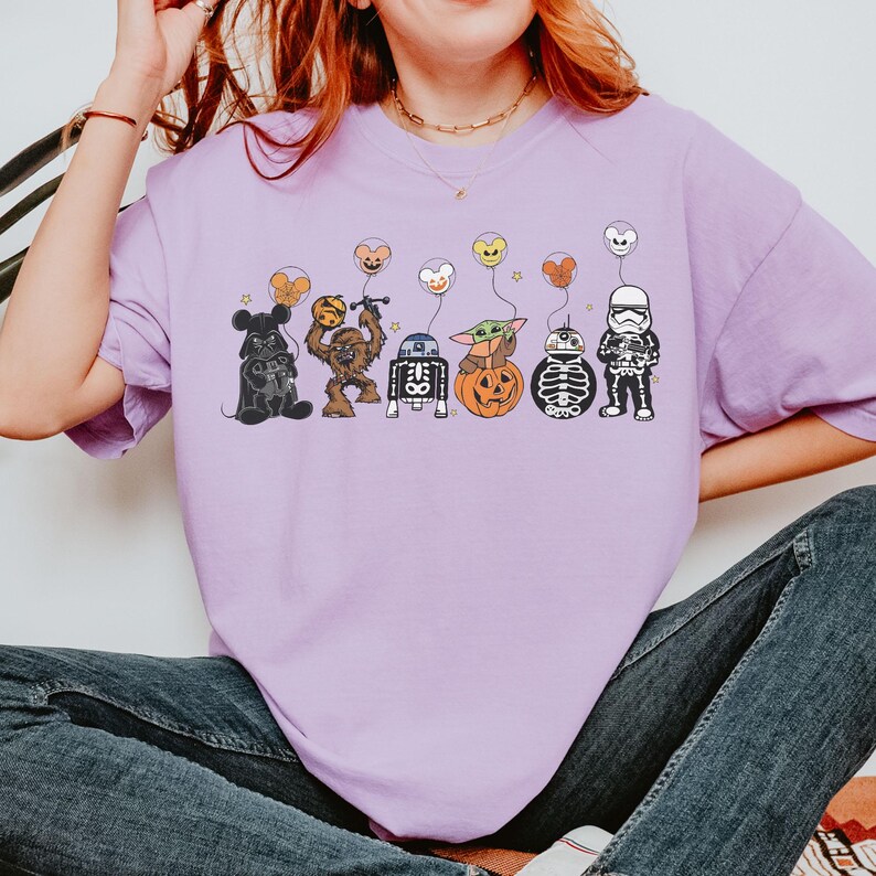Puede incluir: Una camiseta de color lila claro con personajes de dibujos animados de Star Wars y otras franquicias, cada uno sosteniendo un globo en forma de coraz&oacute;n. Los personajes incluyen Darth Vader, Chewbacca, R2-D2, Baby Yoda, un droide esqueleto y un Stormtrooper.