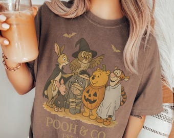Puede incluir: Una camiseta marrón de manga corta con un gráfico de los personajes de Winnie the Pooh con disfraces de Halloween, incluyendo una calabaza, un fantasma y una momia. El texto dice POOH & CO. EST. 1926, junto a siluetas de murciélagos.