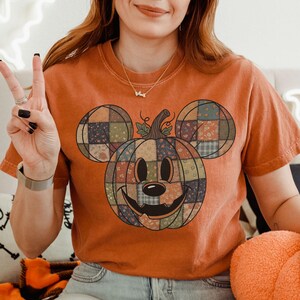 May include: Orange t-shirt featuring a patchwork pumpkin design in the shape of a mouse. The pumpkin has a smiling face and is made of various fabric patterns. The shirt is short-sleeved and has a crew neck.