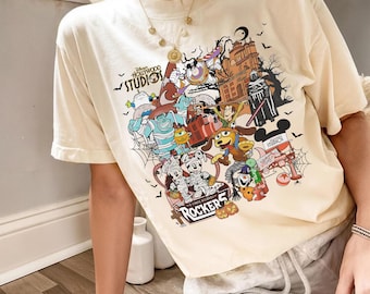 Puede incluir: Camiseta color crema con un gráfico colorido de personajes de Disney y el texto "Hollywood Studios 31". El diseño incluye figuras de dibujos animados, una casa embrujada y la palabra "Rocker".