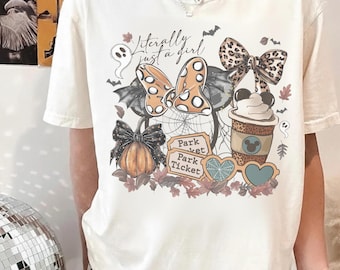 Puede incluir: Una camiseta de manga corta color crema con un estampado gráfico que incluye el texto "Literally just a girl". El diseño muestra una calabaza, una taza de café, gafas en forma de corazón, entradas de parque, un lazo de leopardo y orejas de ratón con alas de murciélago.