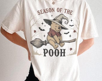 Puede incluir: Camiseta color crema con un diseño de Winnie the Pooh disfrazado de bruja, montado en una escoba. El texto "SEASON OF THE POOH" está arqueado arriba y abajo de la imagen, con murciélagos y estrellas.