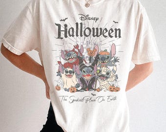 Puede incluir: Una camiseta de manga corta de color crema con un gráfico de Disney Halloween. El estampado muestra varios personajes de Stitch con diversos disfraces, incluyendo una momia y un diablo, con el texto Disney Halloween y The Spookiest Place On Earth.