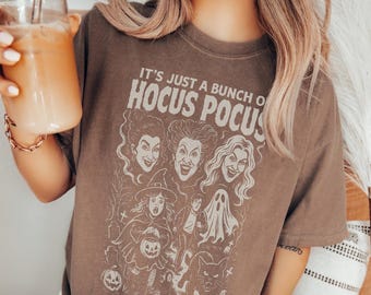 Puede incluir: Una camiseta marrón de manga corta con un estampado gráfico color crema con el texto IT'S JUST A BUNCH OF HOCUS POCUS. El diseño incluye ilustraciones de tres mujeres, un niño disfrazado, un fantasma, un gato y una calabaza en un cementerio.
