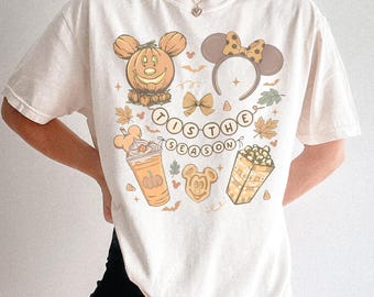 Puede incluir: Camiseta color crema con ilustraciones temáticas de otoño. El diseño incluye una calabaza con orejas de Mickey Mouse, una diadema con orejas de Minnie Mouse y la frase "'Tis the Season". Otros gráficos incluyen hojas de otoño, una bebida de café y palomitas de maíz.