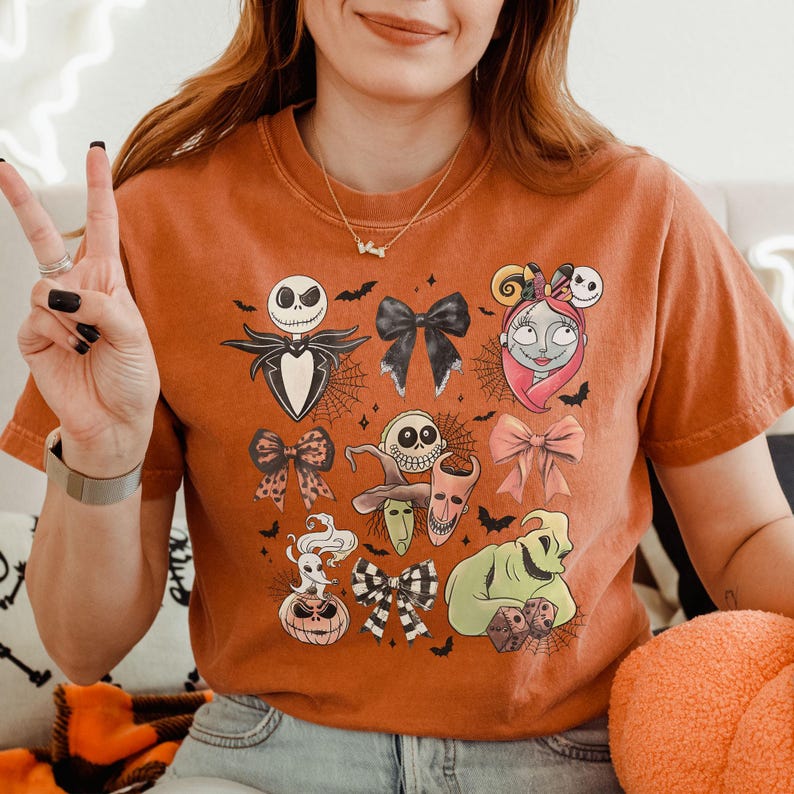 K&ouml;nnte beinhalten: Ein orangefarbenes T-Shirt mit Halloween-Motiven und Schleifendesigns. Die Grafik zeigt Jack Skellington, Sally und Oogie Boogie, mit Spinnweben und Flederm&auml;usen. Das T-Shirt hat kurze &Auml;rmel.