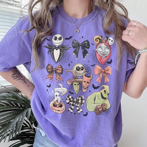K&ouml;nnte beinhalten: Ein lila T-Shirt mit einem Design mit Charakteren und Schleifen aus "The Nightmare Before Christmas". Die Grafik zeigt Jack Skellington, Sally und Oogie Boogie mit Spinnweben und Flederm&auml;usen. Das T-Shirt hat einen Rundhalsausschnitt und kurze &Auml;rmel.