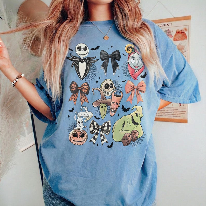 K&ouml;nnte beinhalten: Hellblaues T-Shirt mit verschiedenen Charakteren und Schleifendesigns aus dem Film "Nightmare Before Christmas". Das Shirt zeigt Bilder von Jack Skellington, Sally und Oogie Boogie sowie Spinnweben und Flederm&auml;usen.