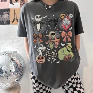 K&ouml;nnte beinhalten: Dunkelgraues T-Shirt mit einer farbenfrohen Grafik von Charakteren und Schleifen aus The Nightmare Before Christmas. Das Design umfasst Jack Skellington, Sally und Oogie Boogie. Das Shirt ist kurz&auml;rmlig und hat einen Rundhalsausschnitt.