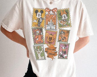 Puede incluir: Una camiseta de manga corta color hueso con un estampado gráfico de ilustraciones estilo sello postal de personajes de dibujos animados con disfraces de Halloween, un castillo y una calabaza, con una pancarta que dice Trick or Treat.