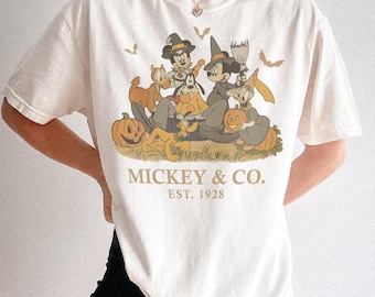 Puede incluir: Una camiseta de manga corta color crema con un gráfico de estilo vintage de personajes de Disney con disfraces de Halloween, incluyendo calabazas y murciélagos. El texto MICKEY & CO. EST. 1928 aparece en una fuente serif color canela.
