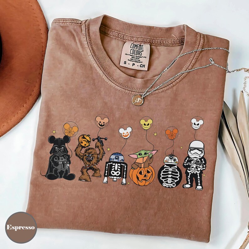 Puede incluir: Camiseta color espresso con personajes de dibujos animados disfrazados para Halloween, incluyendo Darth Vader, Chewbacca, R2-D2, Baby Yoda y un Stormtrooper. La camiseta tiene la etiqueta de la marca Comfort Colors.