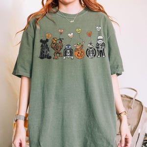 Puede incluir: Camiseta verde oliva con un dise&ntilde;o tem&aacute;tico de Halloween. El dise&ntilde;o incluye personajes de dibujos animados como Mickey Mouse, Chewbacca, R2-D2, Baby Yoda, una calabaza y un Stormtrooper, cada uno sosteniendo un globo. La camiseta tiene un corte holgado.