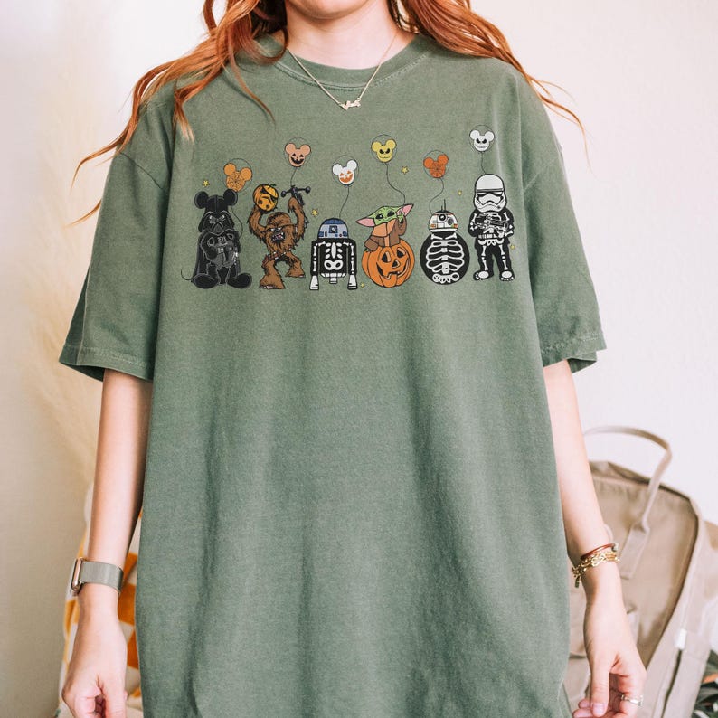 Puede incluir: Camiseta verde oliva con un dise&ntilde;o tem&aacute;tico de Halloween. El dise&ntilde;o incluye personajes de dibujos animados como Mickey Mouse, Chewbacca, R2-D2, Baby Yoda, una calabaza y un Stormtrooper, cada uno sosteniendo un globo. La camiseta tiene un corte holgado.