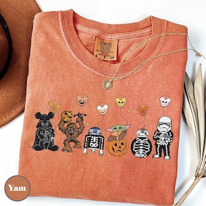Puede incluir: Camiseta naranja Comfort Colors con personajes de Star Wars disfrazados para Halloween. Darth Vader, Chewbacca, R2-D2, Baby Yoda, BB-8 y un Stormtrooper se muestran con globos de Mickey Mouse. Un collar dorado descansa sobre la camiseta.