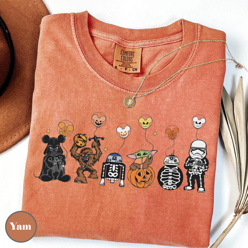 Puede incluir: Camiseta naranja Comfort Colors con personajes de Star Wars disfrazados para Halloween. Darth Vader, Chewbacca, R2-D2, Baby Yoda, BB-8 y un Stormtrooper se muestran con globos de Mickey Mouse. Un collar dorado descansa sobre la camiseta.