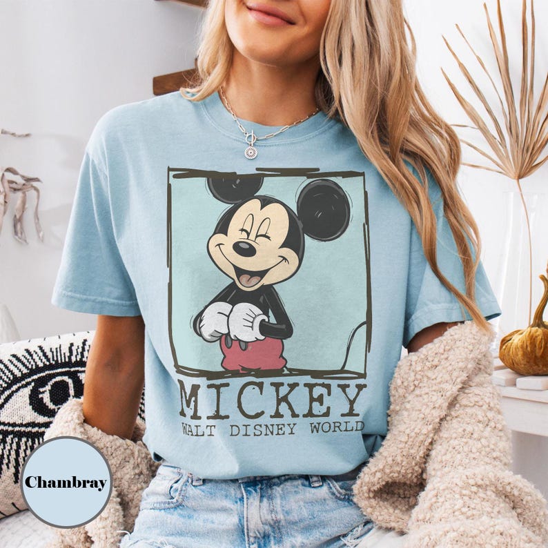 May include: Pale blue t-shirt with a Mickey Mouse graphic in a square frame. The graphic includes the text "MICKEY WALT DISNEY WORLD". The shirt is a solid colour and has short sleeves. The design has a vintage style.