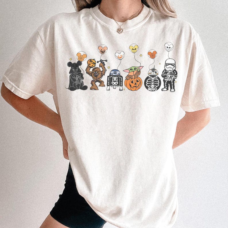 Puede incluir: Camiseta color crema con un dise&ntilde;o de Star Wars con tem&aacute;tica de Halloween. El dise&ntilde;o incluye a Darth Vader, Chewbacca, R2-D2, Baby Yoda en una calabaza, un esqueleto y un Stormtrooper, cada uno sosteniendo un globo.