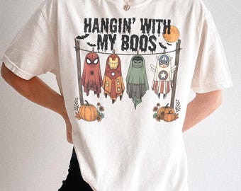 Puede incluir: Camiseta color crema con el texto "HANGIN' WITH MY BOOS" sobre fantasmas de superhéroes de dibujos animados. Los fantasmas están disfrazados de Spider-Man, Iron Man, Hulk y Capitán América. También se representan calabazas y una luna llena.