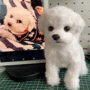May include: A white plush dog figurine with a textured, fluffy coat, dark round eyes, and a brown nose. The figurine is standing upright, featuring realistic facial features and soft fur, positioned next to a screen displaying a similar white dog.