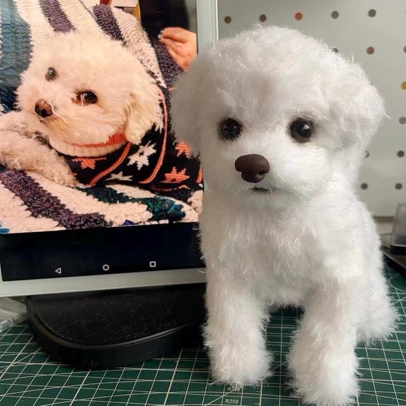 May include: A white plush dog figurine with a textured, fluffy coat, dark round eyes, and a brown nose. The figurine is standing upright, featuring realistic facial features and soft fur, positioned next to a screen displaying a similar white dog.
