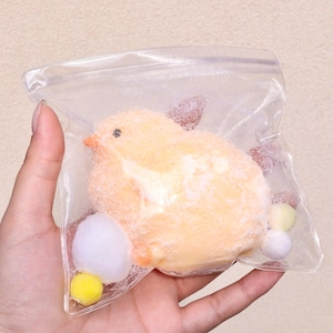 Puede incluir: Un juguete de peluche amarillo en forma de pollito en una bolsa de plástico transparente con pompones blancos y amarillos.