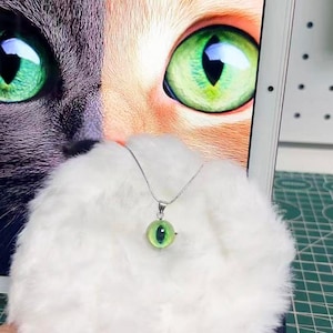 Puede incluir: Un collar de tono plateado con un colgante esférico que presenta un diseño de ojo de gato verde. El colgante está centrado sobre un material blanco, esponjoso y texturizado, con una imagen grande y detallada de un ojo de gato verde visible al fondo.
