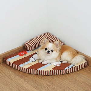 May include: A small, fluffy, tan and white dog rests on a corner pet bed. The bed has a striped brown and tan pattern with a matching bolster pillow. A cartoon apple graphic is visible on the bed.