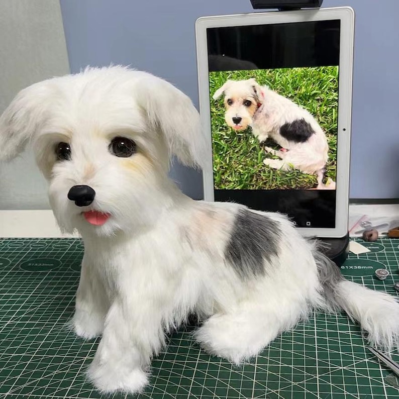 May include: A plush dog figurine with white fur, a black patch on its back, dark eyes, a black nose, and a pink tongue. Behind the figurine, a tablet displays a photograph of a similar dog sitting on green grass, mirroring the plush toy's appearance.
