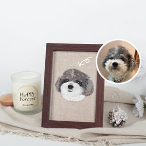 Puede incluir: Retrato bordado de perro en un marco de madera marrón oscuro. La obra presenta un perro gris y blanco sobre un fondo blanco. A la izquierda del marco hay una vela con el texto "Happy Forever".