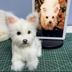May include: A white plush dog figurine with pointed ears, dark eyes, and a brown nose. Beside the figurine stands a digital tablet displaying a photograph of the same white dog. The plush toy features realistic synthetic fur and a seated posture.