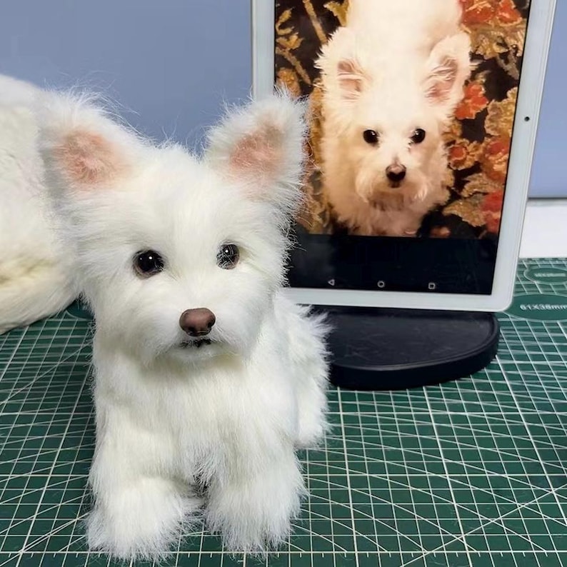 May include: A white plush dog figurine with pointed ears, dark eyes, and a brown nose. Beside the figurine stands a digital tablet displaying a photograph of the same white dog. The plush toy features realistic synthetic fur and a seated posture.