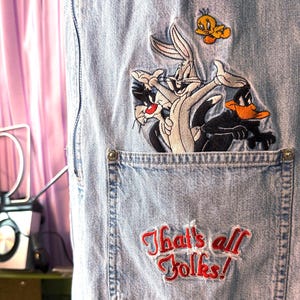 Puede incluir: Mono vaquero azul claro con bordados de los personajes de Looney Tunes: Bugs Bunny, Silvestre, Pato Lucas y Piolín. Un bolsillo muestra la frase "That's all Folks!" en rojo.