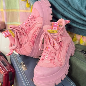 Könnte beinhalten: Ein Paar pinkfarbene Buffalo Plateau-Sneaker. Die High-Top-Schuhe haben dicke, geriffelte Sohlen und sind aus einem strukturierten Material gefertigt. Die Schnürsenkel und die Zunge tragen das Buffalo-Logo. Die Schuhe werden auf einem blauen Koffer präsentiert.
