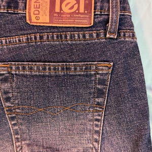 May include: Close-up of the back of blue denim jeans. The jeans feature a back pocket and a brown leather patch with the "lei" logo and "eDENIM" text. The jeans have a classic, worn look.