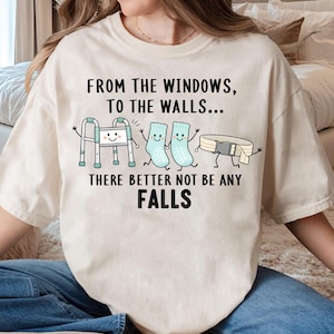 Peut inclure: Un t-shirt à manches courtes couleur crème avec un motif représentant un déambulateur, deux chaussettes et une ceinture de transfert avec des visages. Le texte indique : FROM THE WINDOWS, TO THE WALLS... THERE BETTER NOT BE ANY FALLS.