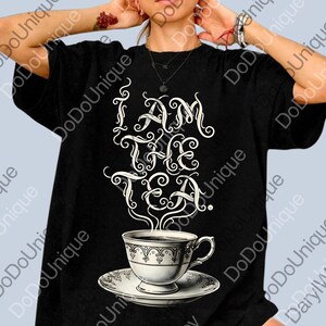 May include: Black t-shirt with the phrase "I AM THE TEA" in elegant white script above a detailed teacup and saucer. The teacup design is white with ornate detailing, and steam rises from the tea.