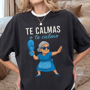 Könnte beinhalten: Schwarzes T-Shirt mit dem Text "TE CALMAS o te calmo" in Weiß und Hellblau. Die Grafik zeigt eine Illustration einer älteren Frau mit Sonnenbrille, die eine blaue Flip-Flop hält, mit einem strengen Gesichtsausdruck und erhobener Hand.