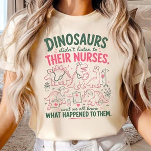Op de afbeelding: Een cr&egrave;mekleurig t-shirt met een afbeelding met de tekst "DINOSAURS didn't listen to THEIR NURSES. and we all know WHAT HAPPENED TO THEM." Het ontwerp bevat illustraties van verschillende dinosaurussen met verpleegstersmutsen en medische hulpmiddelen.