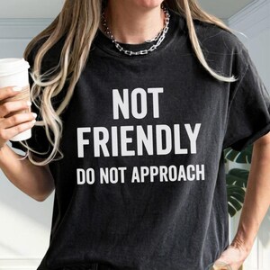 Könnte beinhalten: Ein schwarzes kurzärmeliges T-Shirt mit Rundhalsausschnitt. Auf der Vorderseite befindet sich der weiße Schriftzug NOT FRIENDLY DO NOT APPROACH in einer fetten, serifenlosen Schrift. Der Stoff hat eine leicht verwaschene Optik.