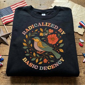 Puede incluir: Sudadera negra con un gráfico colorido que presenta un pájaro, flores y el texto "RADICALIZED BY BASIC DECENCY" en un diseño circular. Una bandera estadounidense y decoraciones patrióticas están en el fondo.