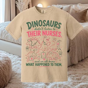 Op de afbeelding: Een beige t-shirt met korte mouwen en een grafische print van dinosaurussen verkleed als verpleegkundigen met medische hulpmiddelen. De tekst luidt: DINOSAURS didn't listen to THEIR NURSES. and we all know WHAT HAPPENED TO THEM. Groene en roze inkt.